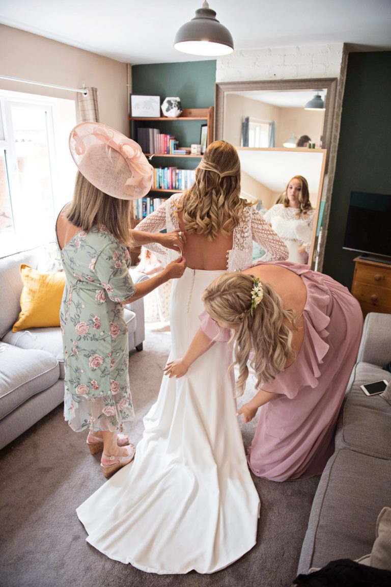 Portrait photo of the bride getting into her dress, helped by the mother of the bride and a bridesmaid.