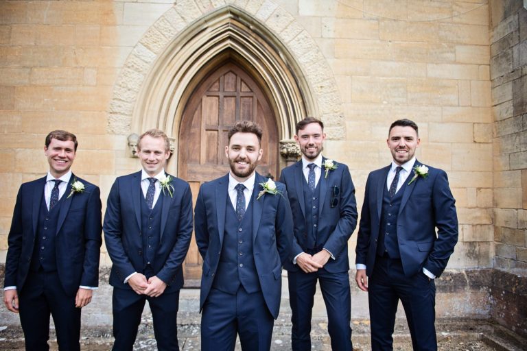 Groomsmen pose outside the church for a photo. All smiling.