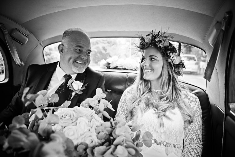 B&W candid photo of bride smiling at her dad in the back of the wedding car.