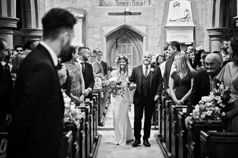B&W candid photo of bride walking down the wedding isle with her dad, watched by wedding guests and groom all smiling.