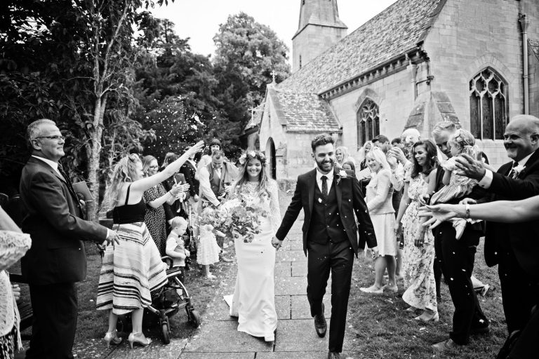 B&W candid image of bride and groom leaving the church as guests throw lots of confetti over them