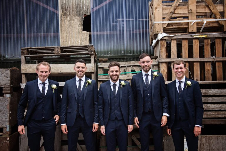 Groomsmen pose in front of wooden pallets. By Blooming Photography