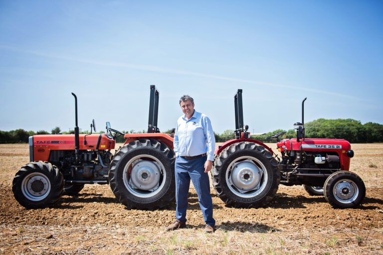 Editorial headshot of a man standing in between two tractors in a field in the UK.