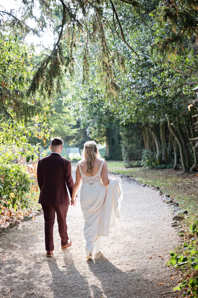 Bride and groom (with their backs to us) walk hand in hand down a woodland gravel path