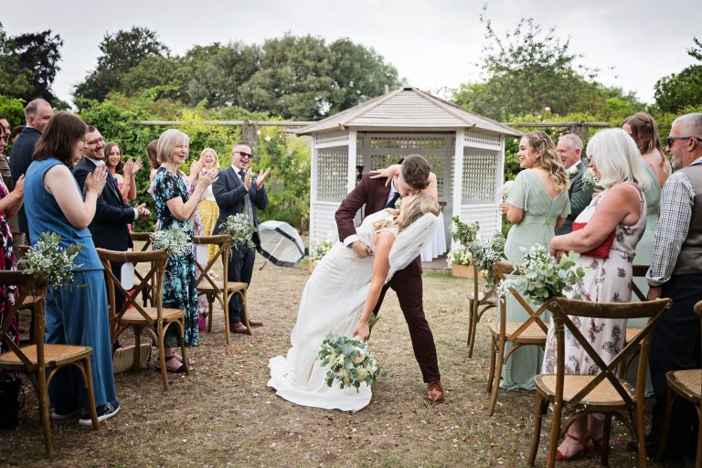 Bride and groom kiss and dip in the middle of the wedding isle with their guests cheering and clapping by them