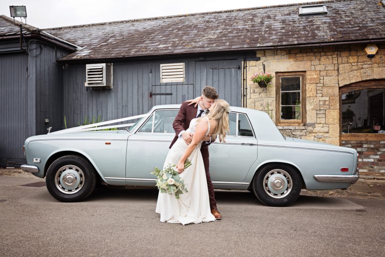 a bride and groom dip and kiss whilst their classic rolls royce is behind them