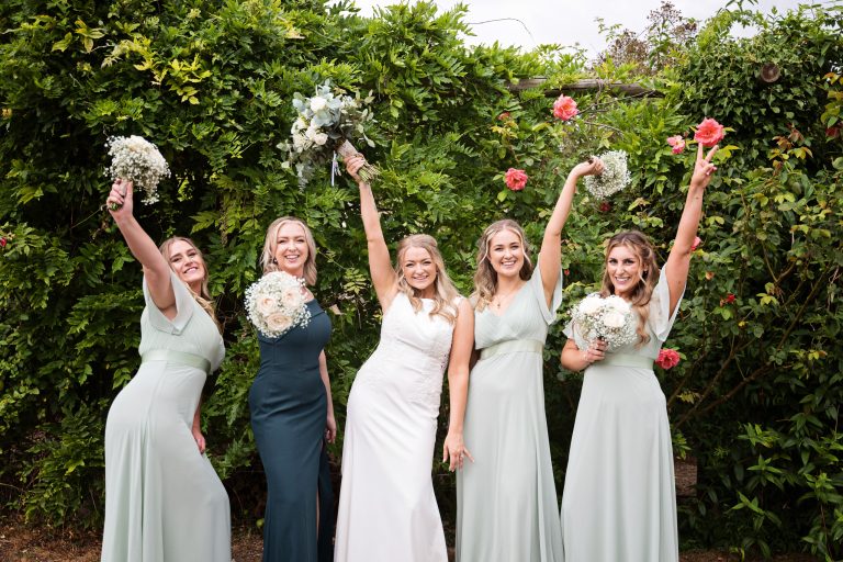 BNridesmaid and bride stand against a floral rose wall with their hands in their air (holding their flower bouquets), smiling