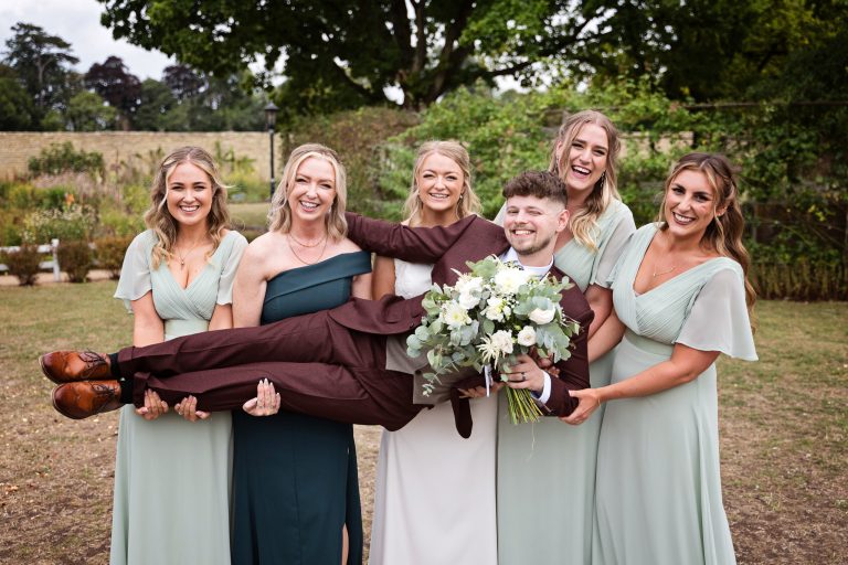 fun image of bridesmaids and bride carrying the groom who is holding the wedding bouquet