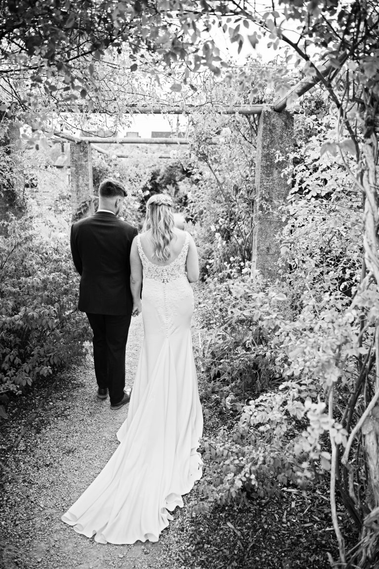 b&w image of bride and groom with their backs to us as they walk through a rose garden. The long train of the brides dress is particularly stunning