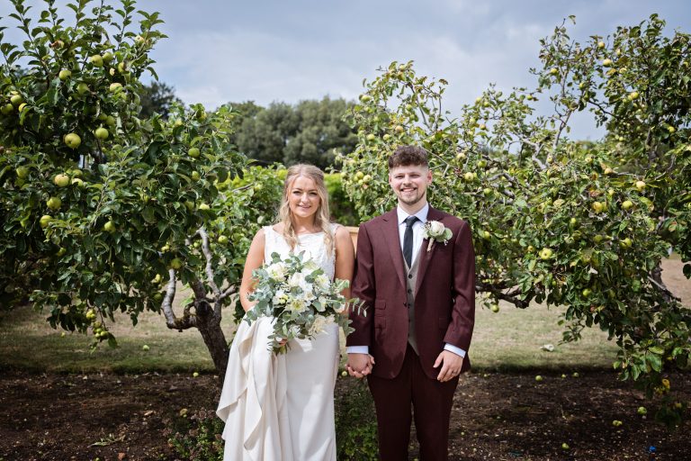 Bride and Groom stand side by side whilst being face on to the camera with apple trees on either side of them.