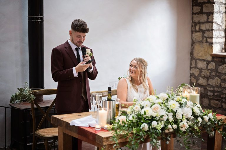groom (standing) next to his bride (who is sitting), as he delivers his wedding speech.