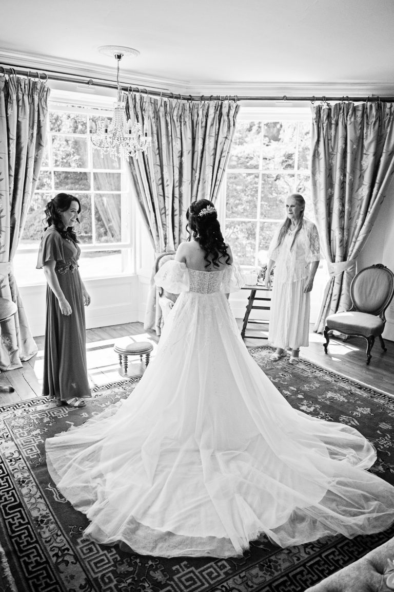 Bridesmaid and mother of the bride emotionally look at the bride in her wedding dress - they are all standing in front of a gorgeous bay window