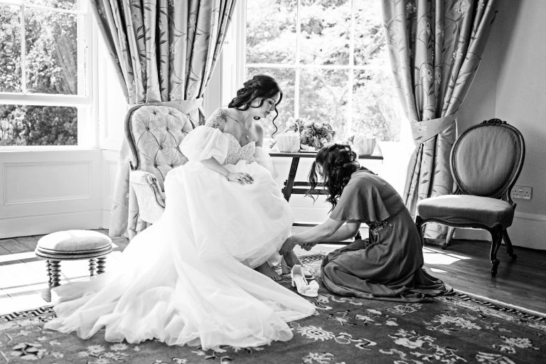 B&W image of a bride sitting having her wedding shoes put on by her sister who is also a bridesmaid. A Georgian bay window is behind them