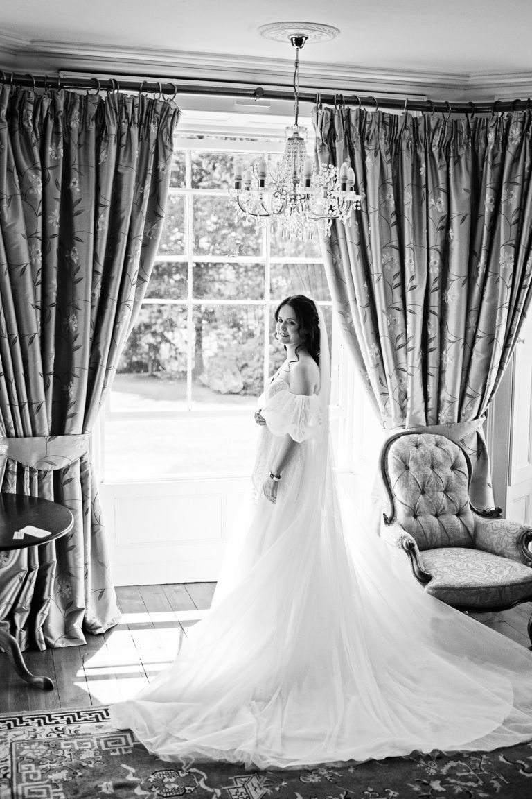 B&W image of a a bride standing in front of a large bay window.