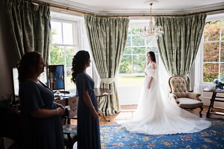 A bride stands in front of a large bay window. Her bridesmaids in the shadows looking at the bride.