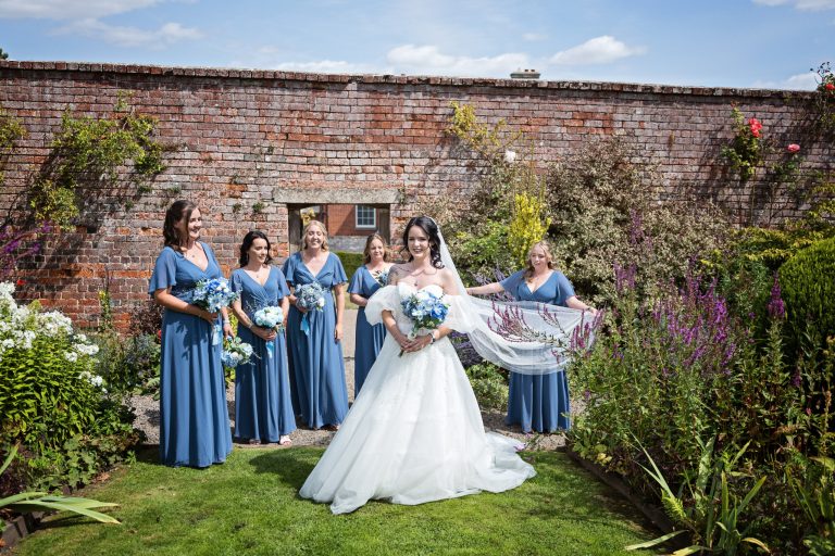 Bridesmaids and bride stand in front of a red brick garden wall with flowers. One bridesmaid is holding out the brides veil.