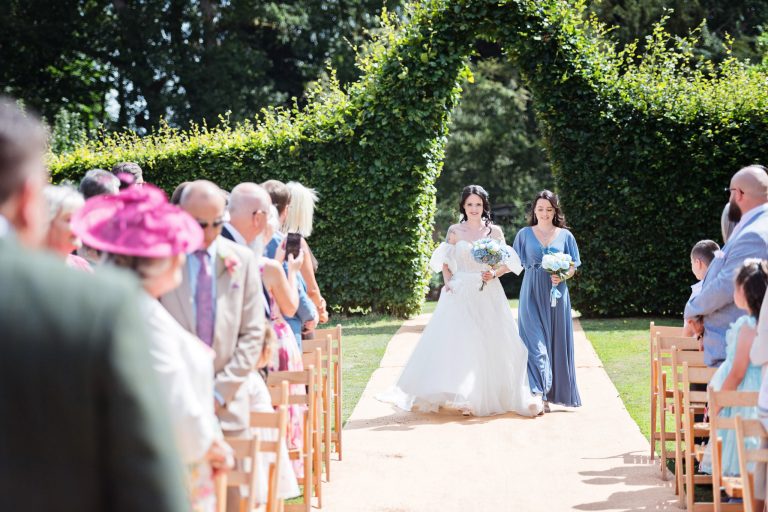 A bride walks down the wedding isle next to her sister. Guests and groom watching