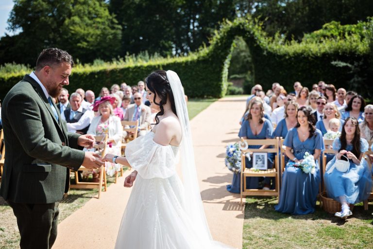 A groom exchanging rings with his bride. Wedding guests smiling in the background
