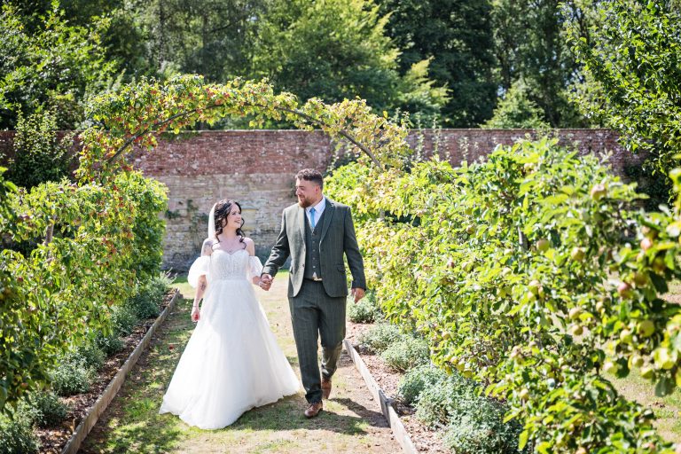 A bride and groom walk hand in hand down an avenue of apple trees