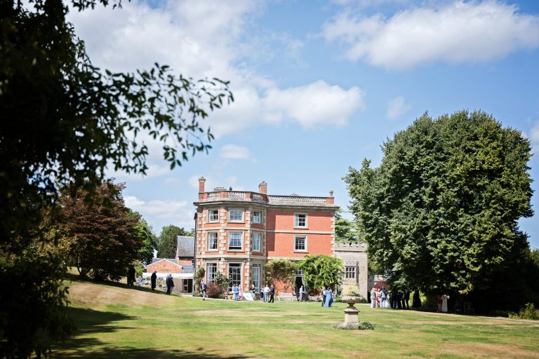 A beautiful English summers day with a Georgian Manor House in frame with a lawn and mature trees surrounding. Blue sky with puffy clouds.