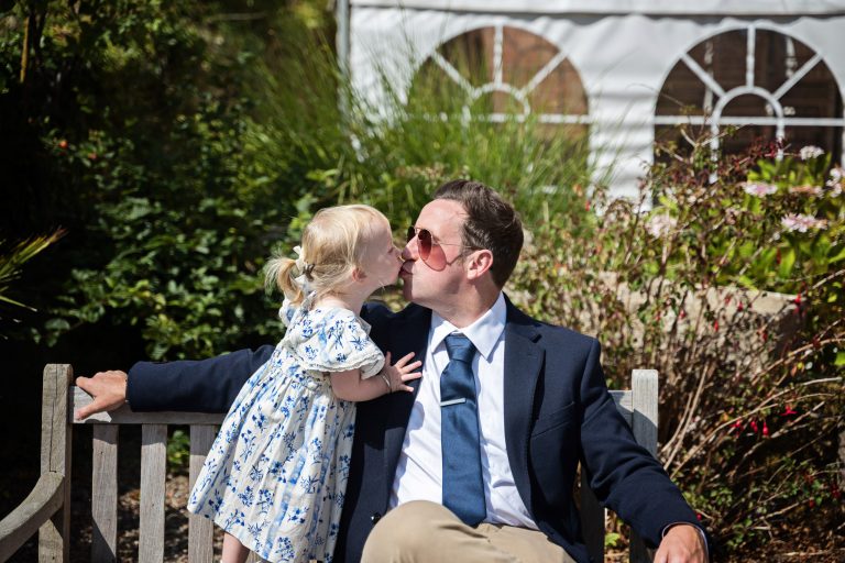 A dad sitting on a bench with sunglasses on being kissed by his daughter