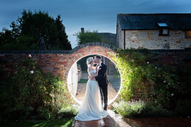 A striking image at dusk of a bride and groom kissing with a red brick wall behind them. They are lit up.