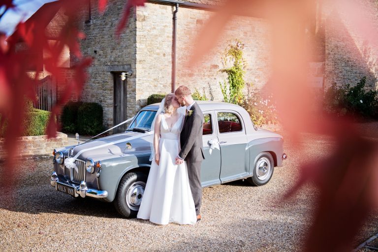 Bride and groom kiss in front of a classic car at Calcot Manor