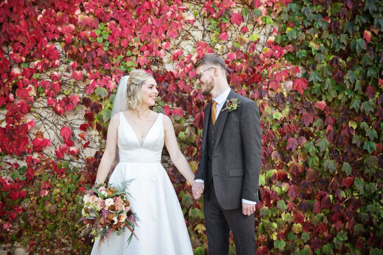 Bride and groom smile and hold hands against a colourful virginia creeper wall