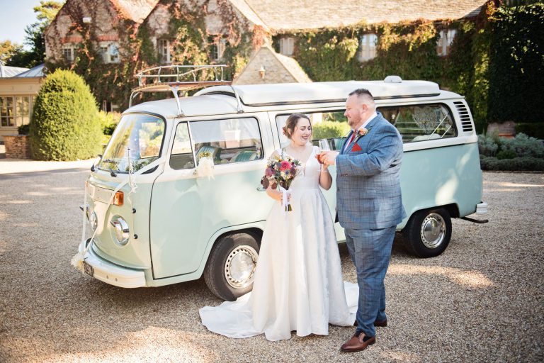 Bride and groom clink their glasses together as they stand in front of a split screen VW outside the front of Calcot Manor