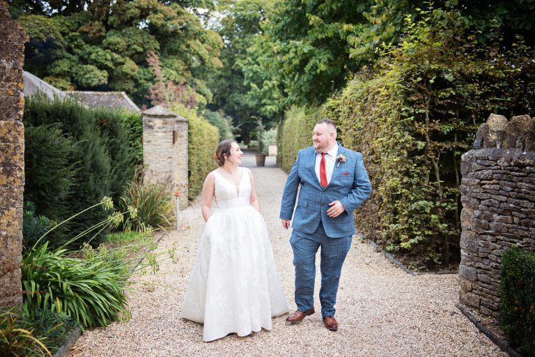 Bride and groom walk down a path at Calcot manor. The bride's hand are in the pockets of her wedding dress.