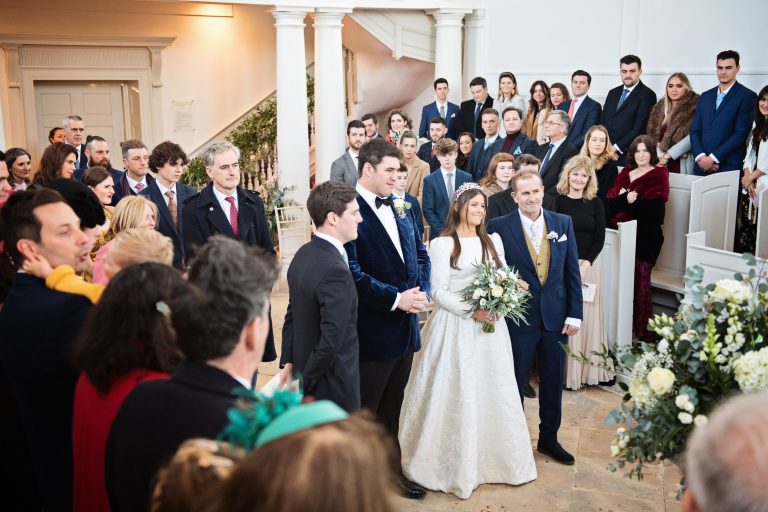 Bride and groom stand with the father of the bride and best man at the start of the wedding ceremony. Wedding guests look onward.