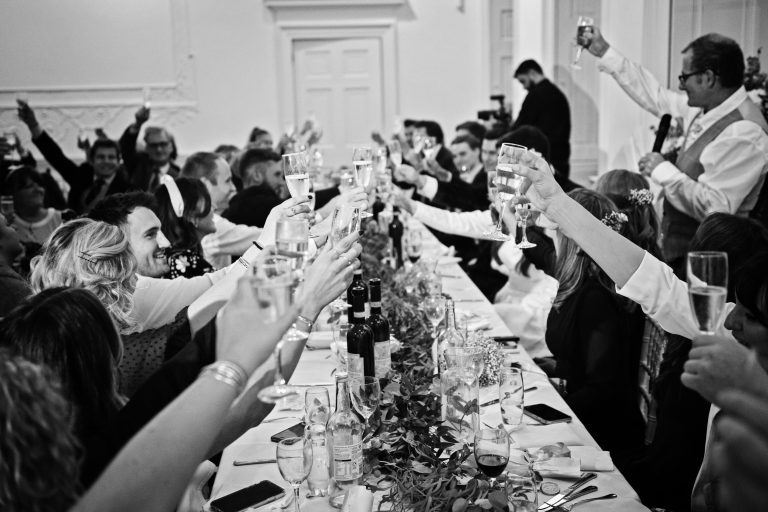 B&W photo of wedding guests raising their glass at the end of father of the brides speech.
