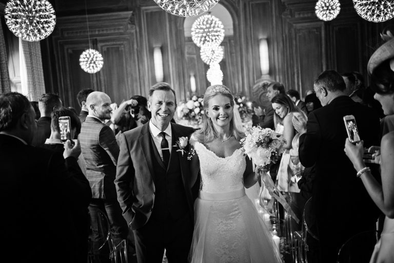 B&W photo of bride and groom walking down the wedding isle after getting married. Friends clapping and taking photos on their phones.