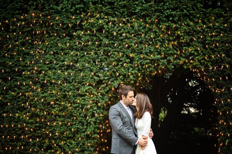 Bride and groom kiss next to a yew hedge with sparkly lights.
