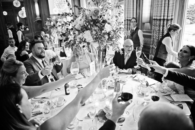 B&W candid photo of wedding guests at Cowley Manor celebrating by raising their glasses to each other whilst they are sat down at a table.