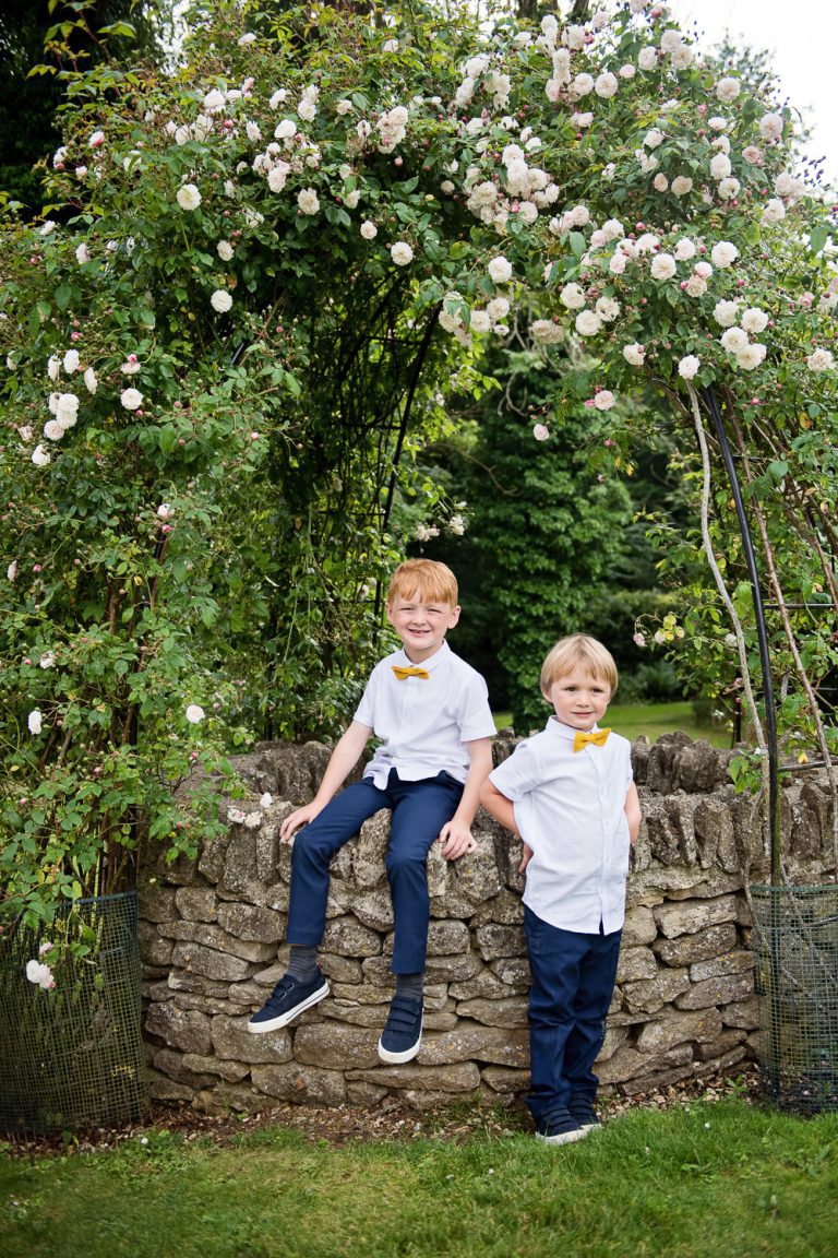 Two cheeky boys stand next to a water well. Beautiful flowering rose frames the image.