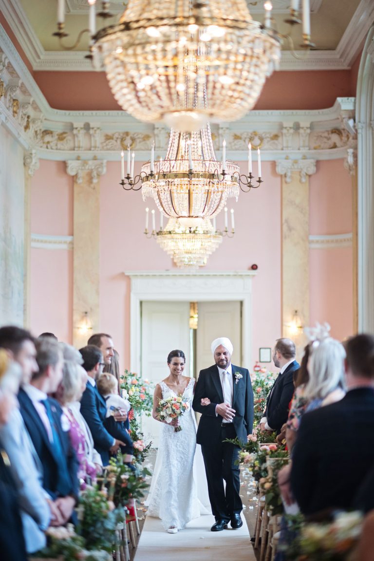 A bride walks down the isle with her father. Chandeliers and ornate fixings make this look luxurious
