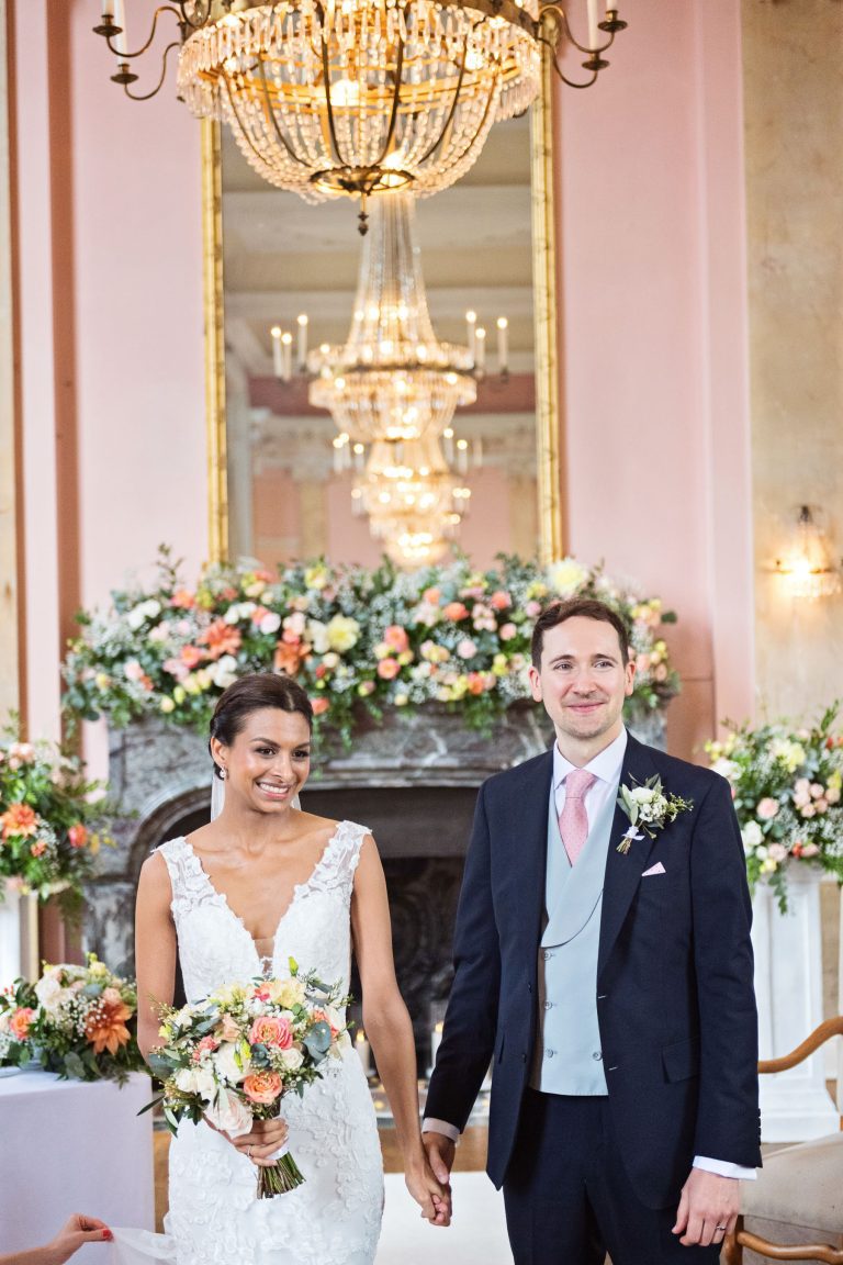 Bride and groom hold hands. Chandeliers and ornate fixings make this look luxurious