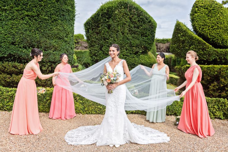 Classic photo of bride standing in the formal grounds at Danesfield House with her bridesmaids laying out her impressive veil.