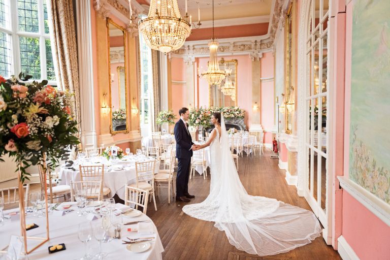 Bride and groom stand in the midst of the wedding breakfast set up at Danesfield House. Chandeliers and ornate fixings make this look luxurious