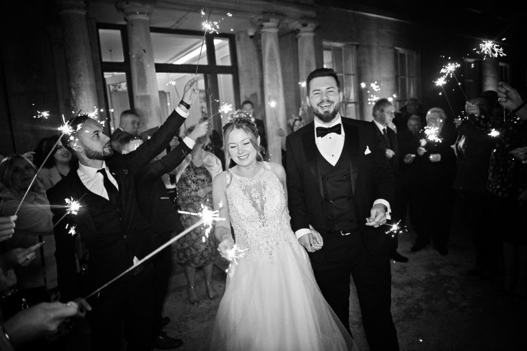 B&W image of Bride and groom with their guests holding sparklers and smiling out the front of Eastington Park.