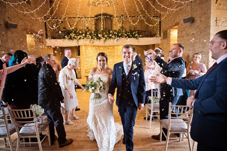 Bride and groom walking down the wedding isle at the Great Tythe Barn with confetti being thrown at them.