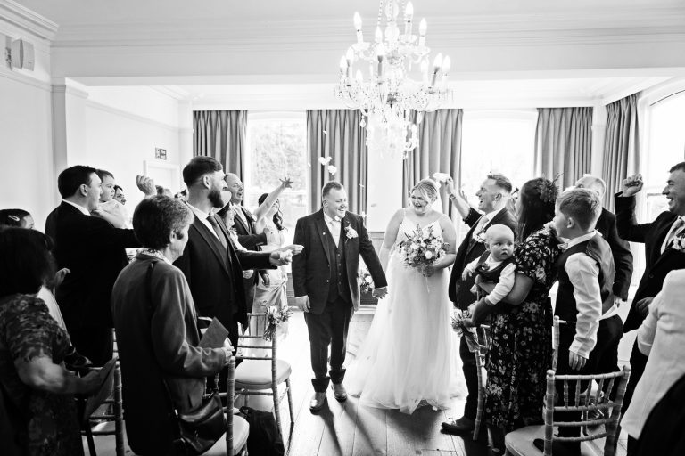 B&W image of bride and groom being showered by confetti as they walk back down the wedding isle at Hatton Court Hotel.