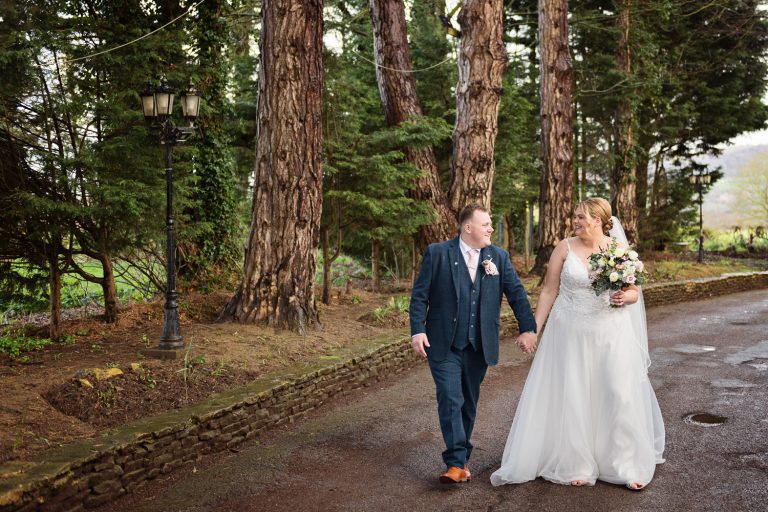 Bride and groom hold hands as they walk down a path. Large pine tree's behind them.