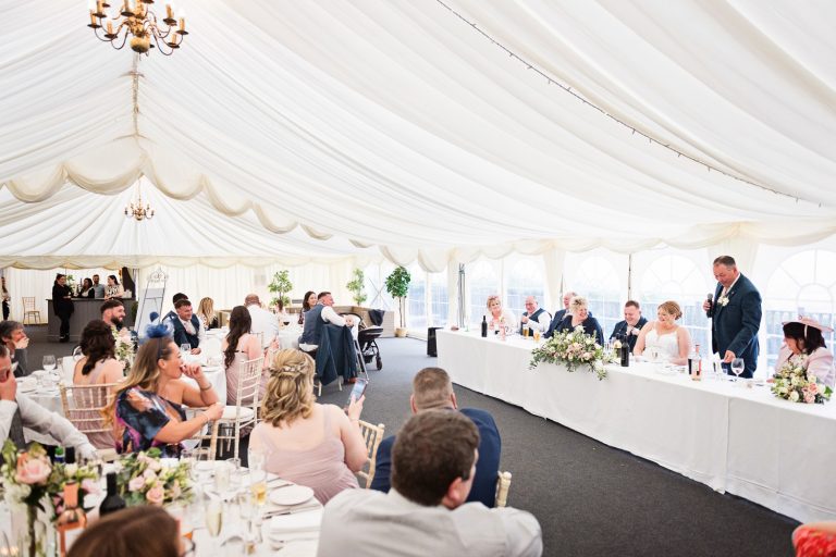 Wedding speech taking place with the father of the bride telling a funny story with wedding guests watching.