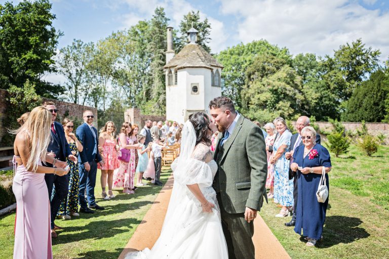 Bride and groom kiss after walking down the wedding isle. Their guests are standing behind them smiling.