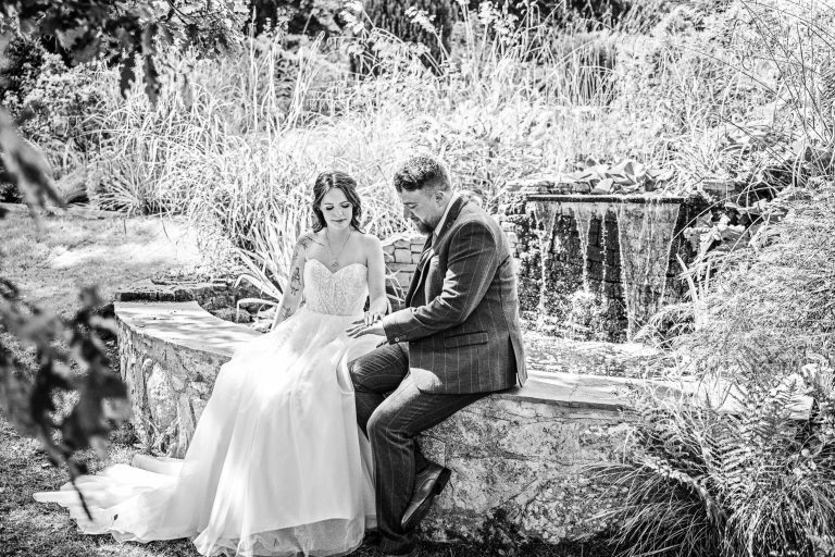 B&W candid photo of bride and groom admiring each others wedding rings whilst they are sat on a brick wall. A small waterfall is behind them.