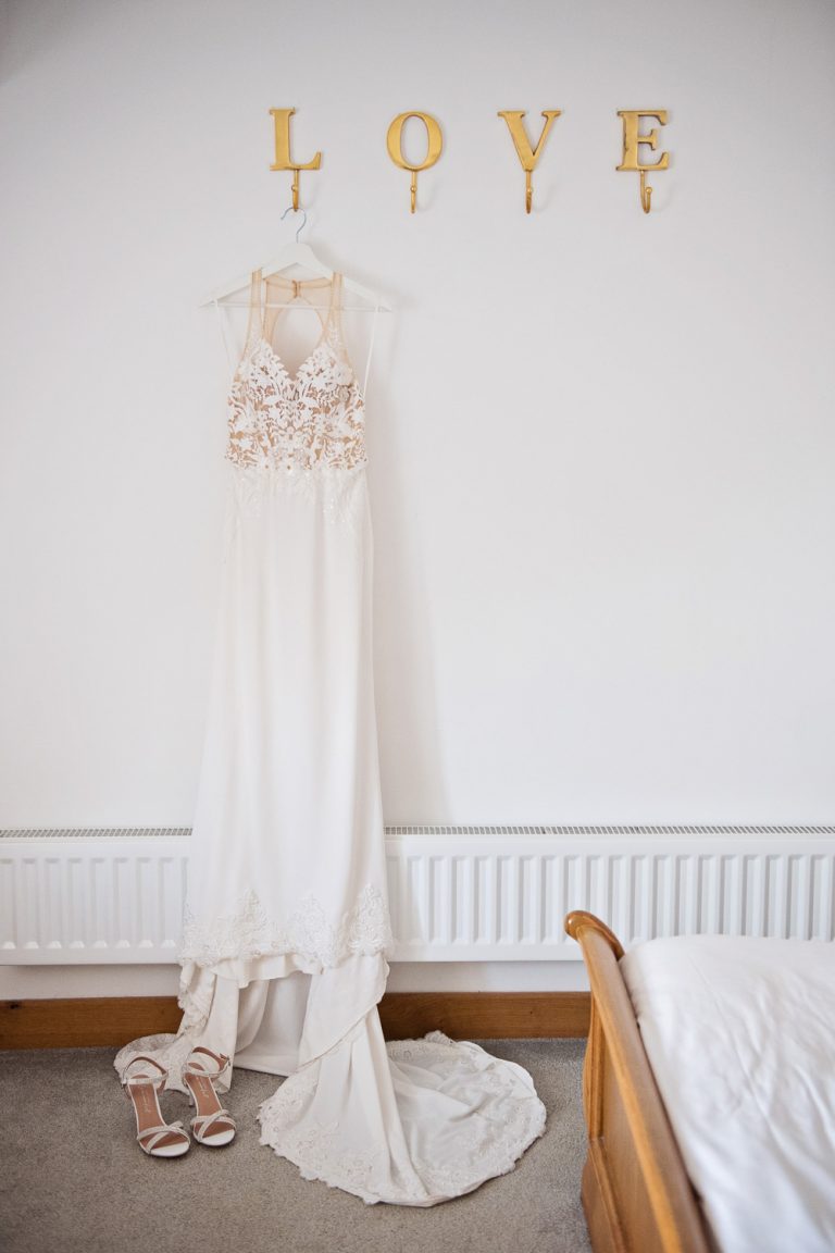 Portrait photo of the dress hung up above "LOVE" letters. Wedding shoes placed below the trail of the dress.
