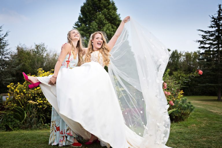A bride shows of her dress and veil for a photo with her friend in the gardens of Huntsmill Farm.