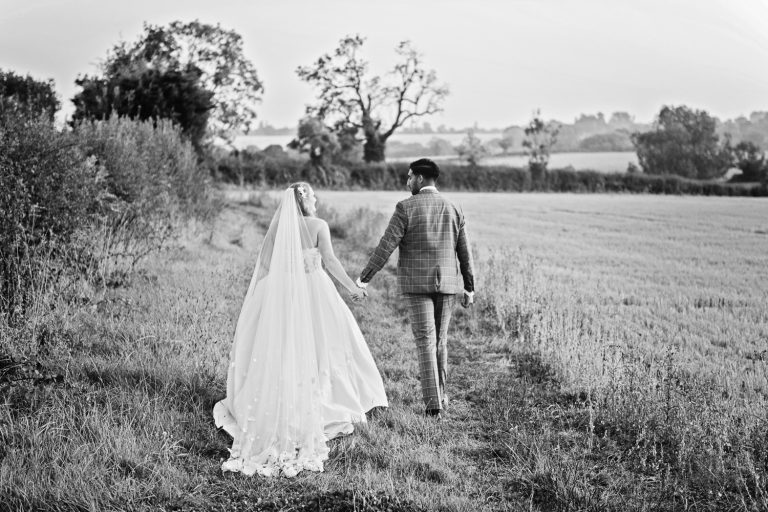 B&W image of Bride and groom walk hand in hand on the edge of a field.